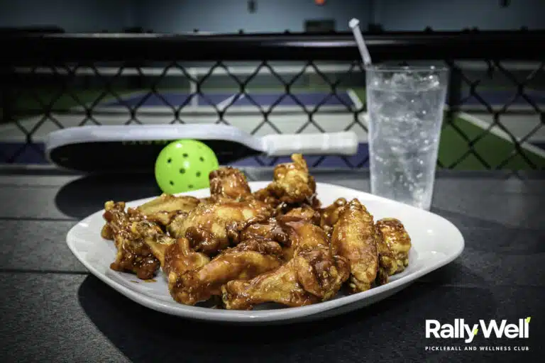 Plate of chicken wings and a drink beside a pickleball paddle and ball on a RallyWell court