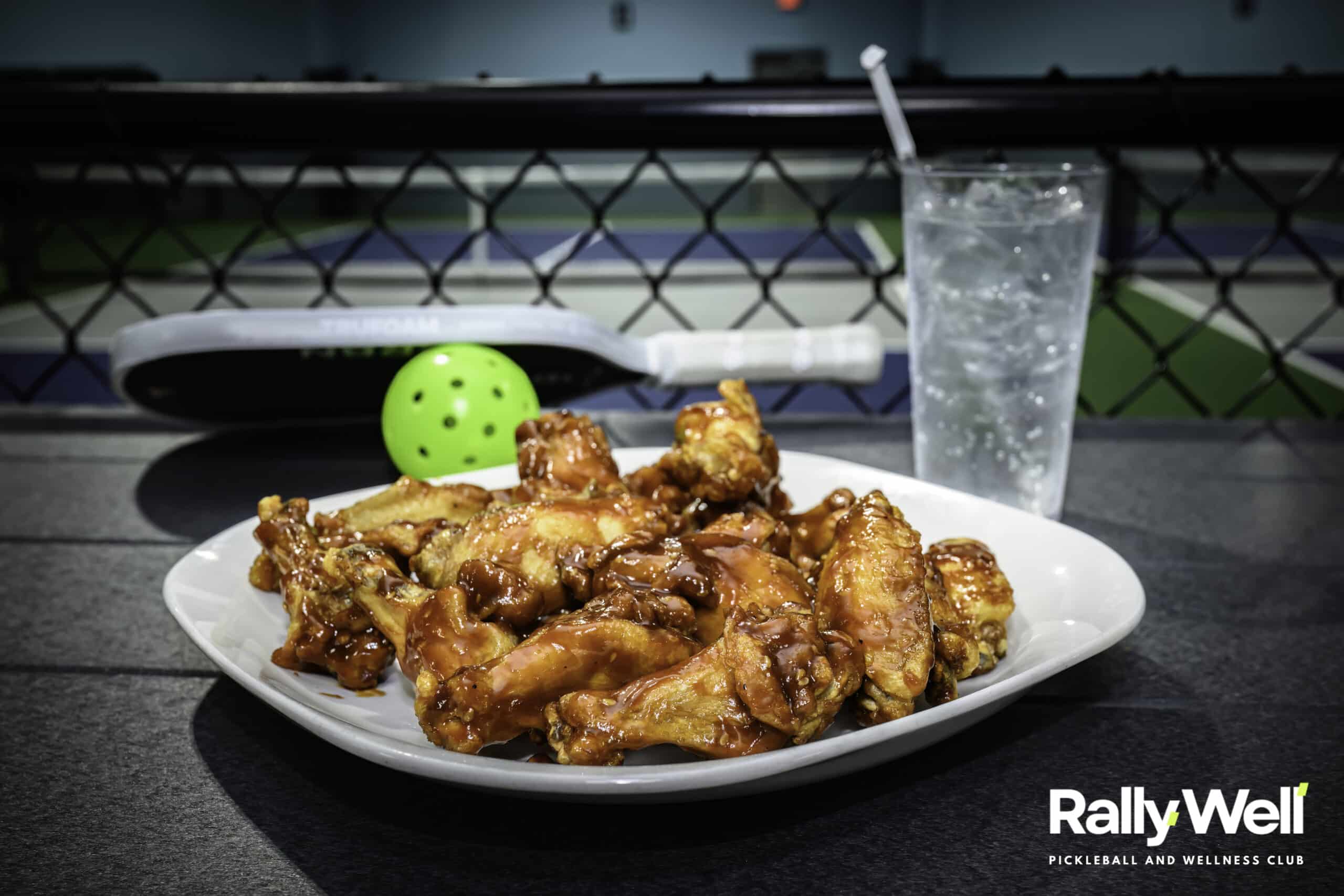 Plate of chicken wings and a drink beside a pickleball paddle and ball on a RallyWell court