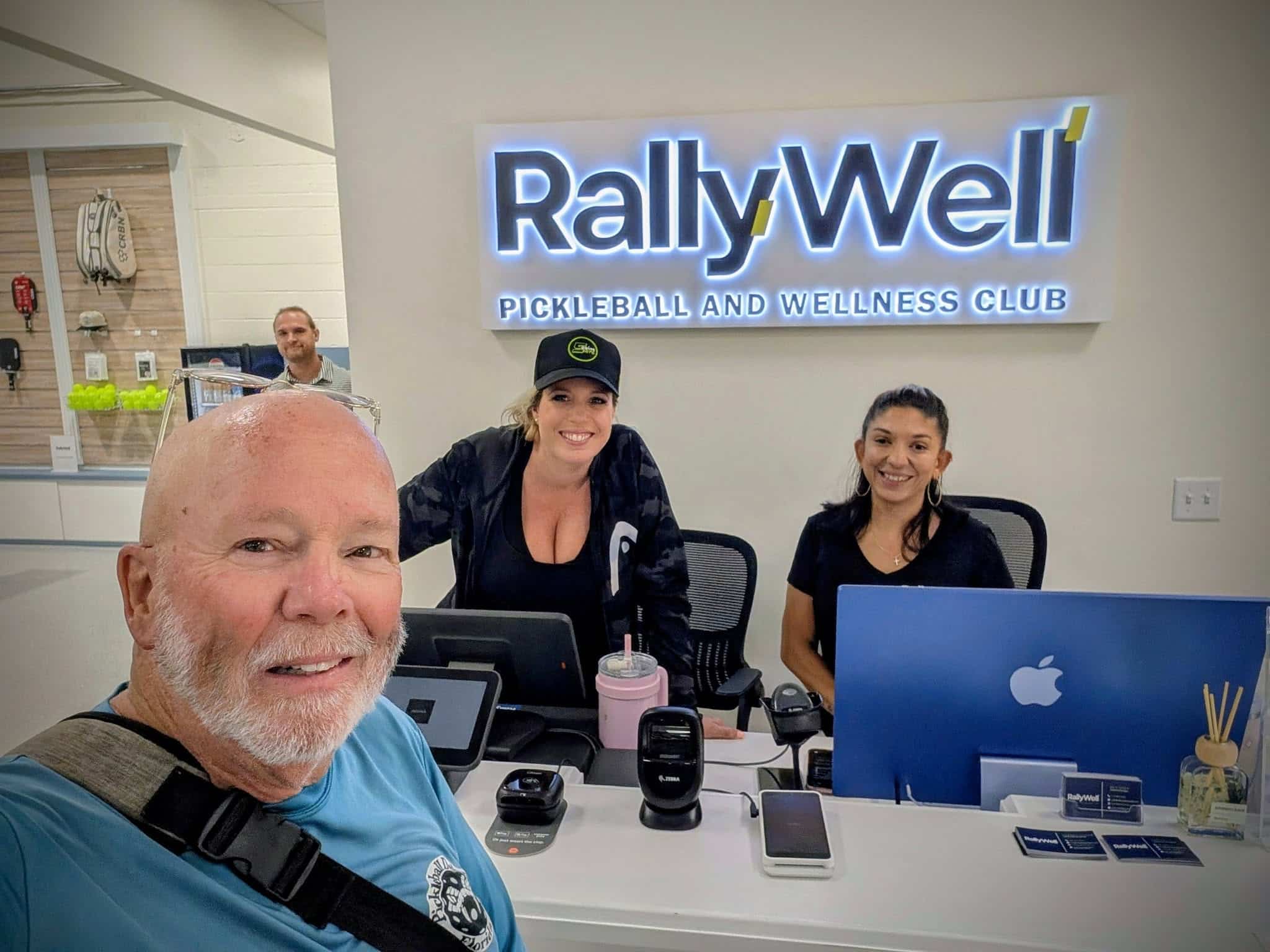 RallyWell front desk staff and a customer with the RallyWell Pickleball and Wellness Club sign